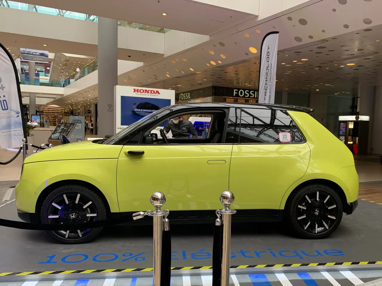 The image shows a yellow car parked in front of a building, surrounded by barrier poles with ropes,...