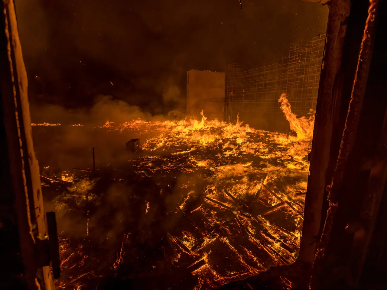 The image shows a raging fire engulfing a building in the middle of a city at night, with smoke...