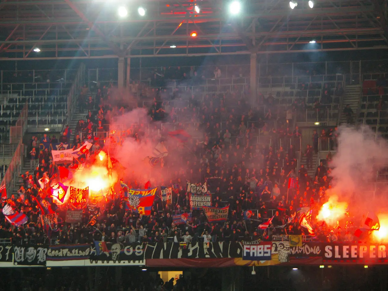 The image shows a large crowd of people in a stadium with flares and smoke billowing out of them....