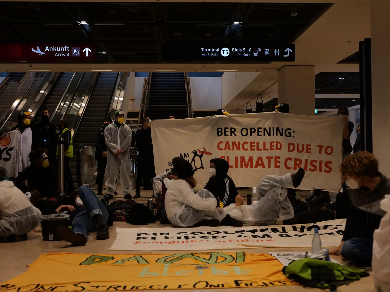 The image shows a group of people sitting on the floor of an airport, holding a banner that reads...