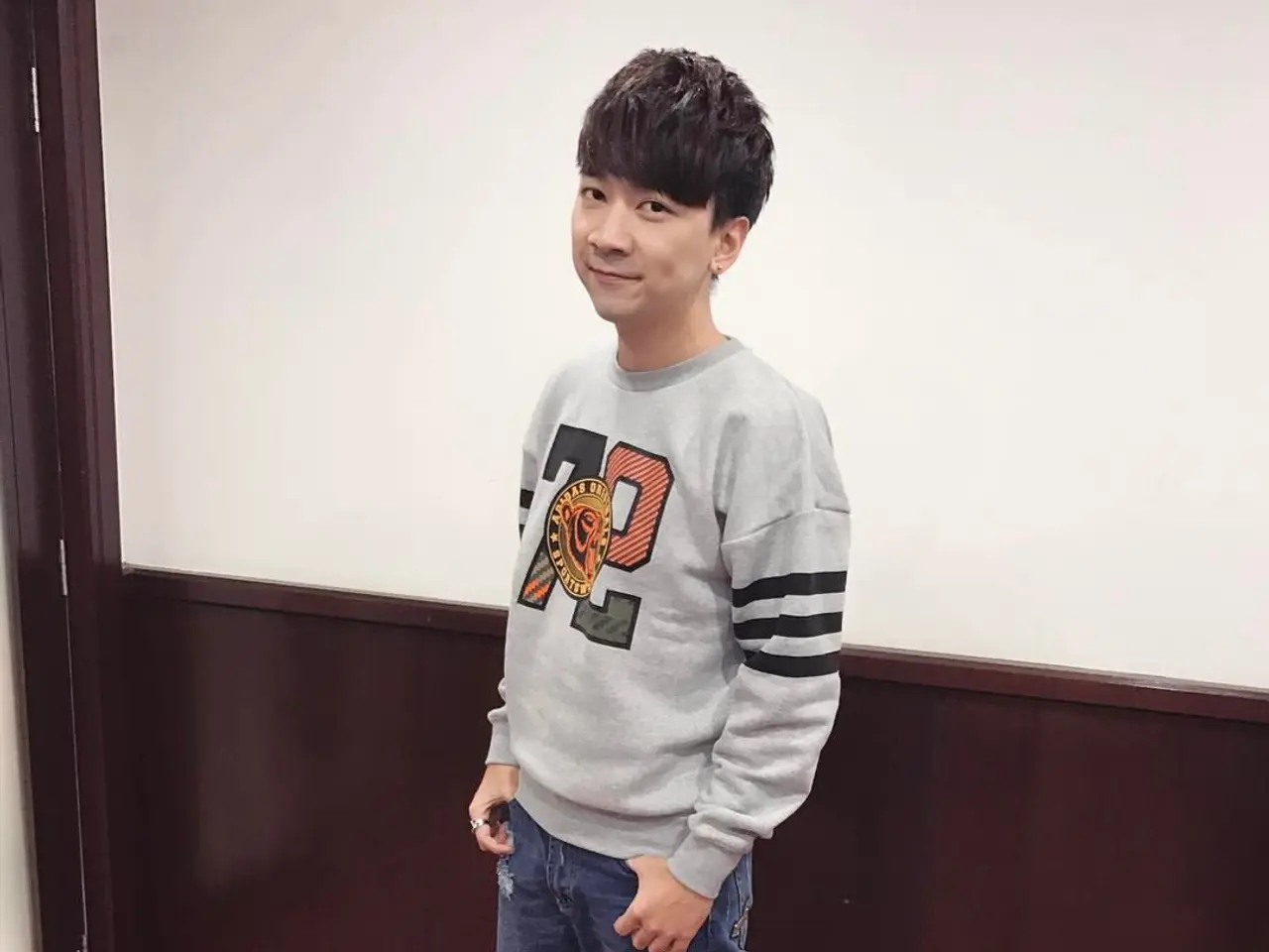 The image shows a young man standing on top of a red carpet, wearing a grey sweatshirt and blue...