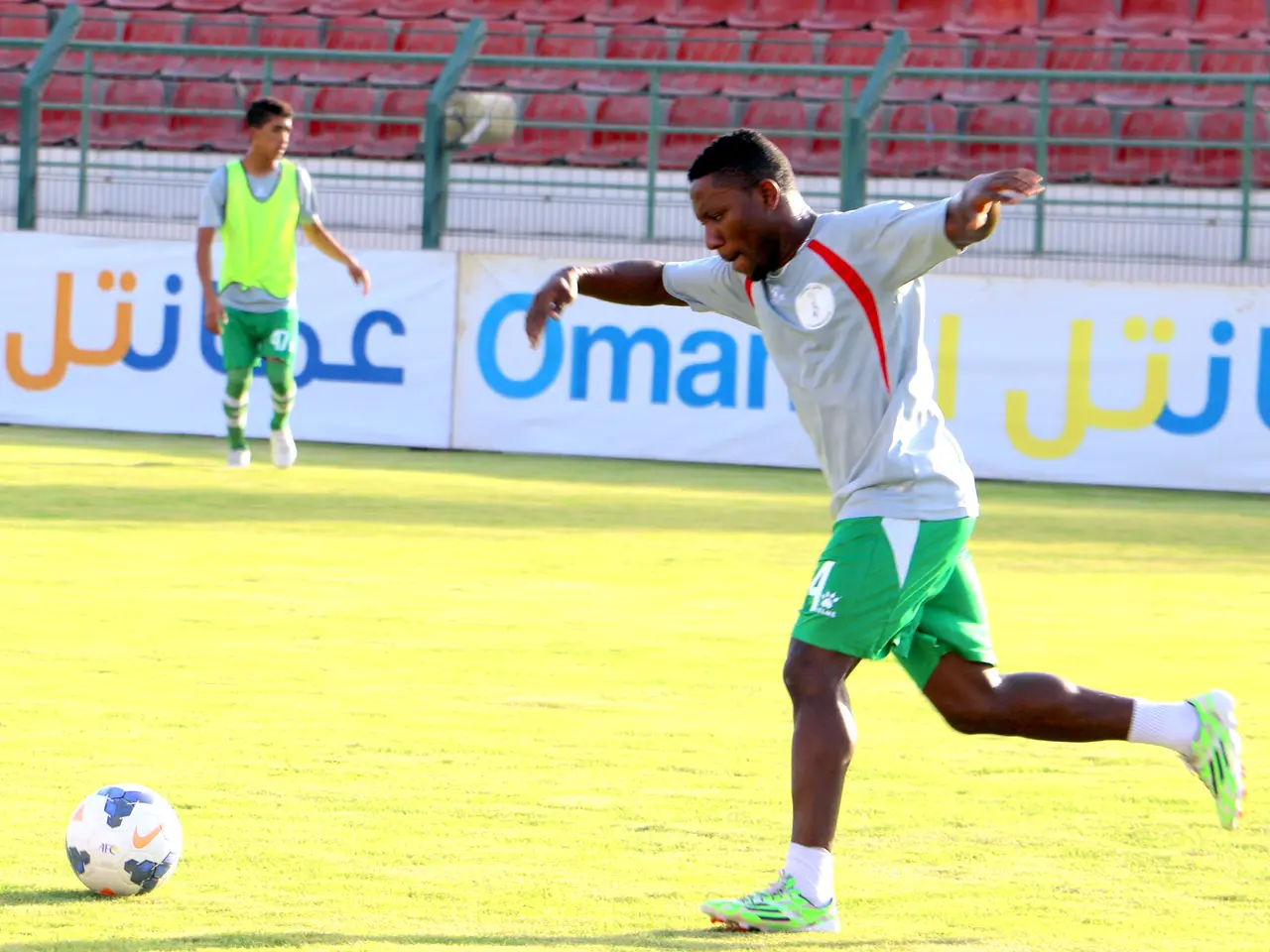 The image shows a man kicking a soccer ball on top of a grassy field, with two other people...