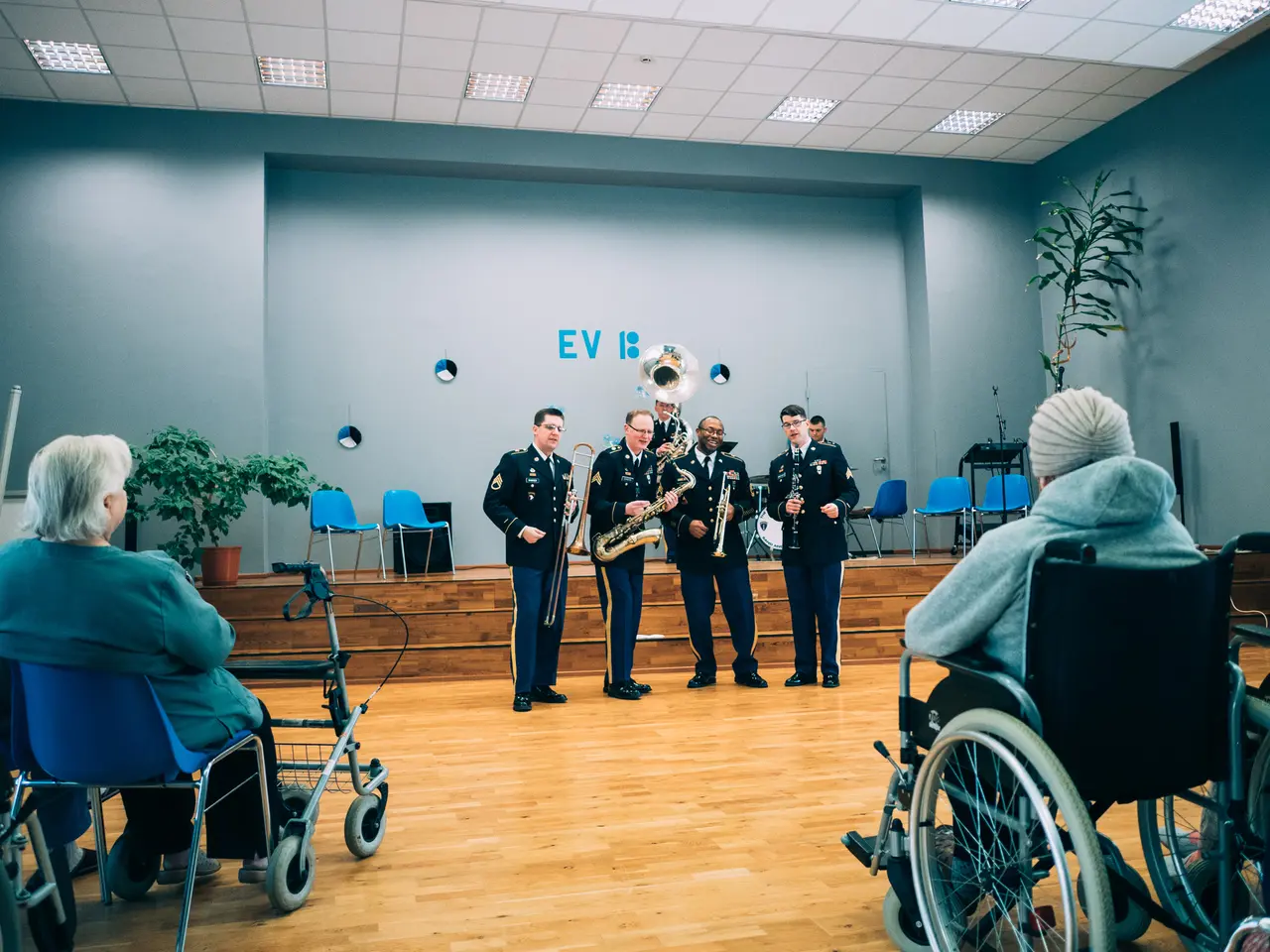 The image shows a group of people in wheelchairs playing musical instruments in a room with a wall...