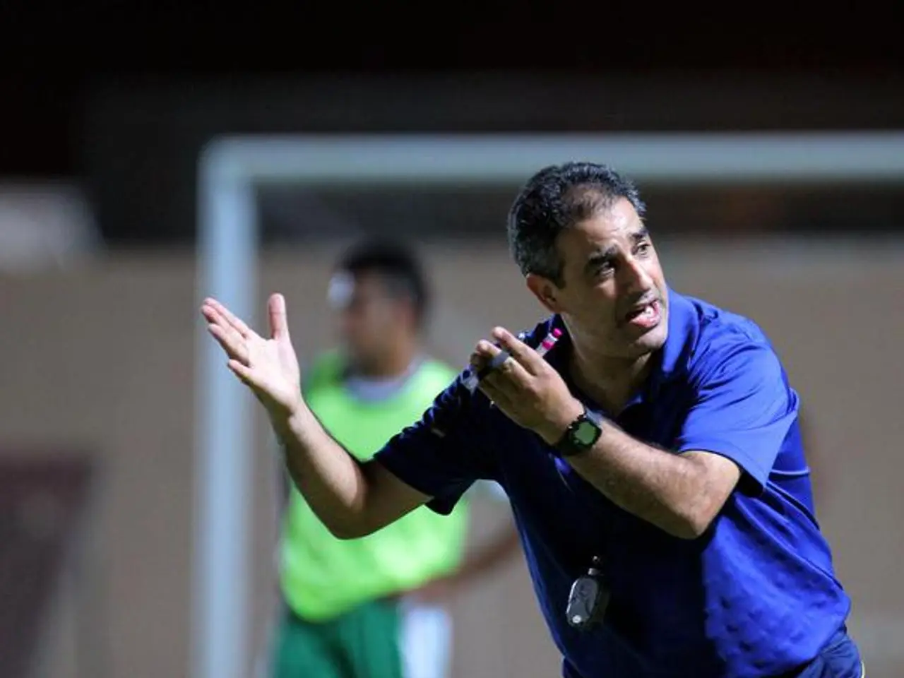 The image shows a man in a blue t-shirt and blue pants standing on a soccer field, holding a...