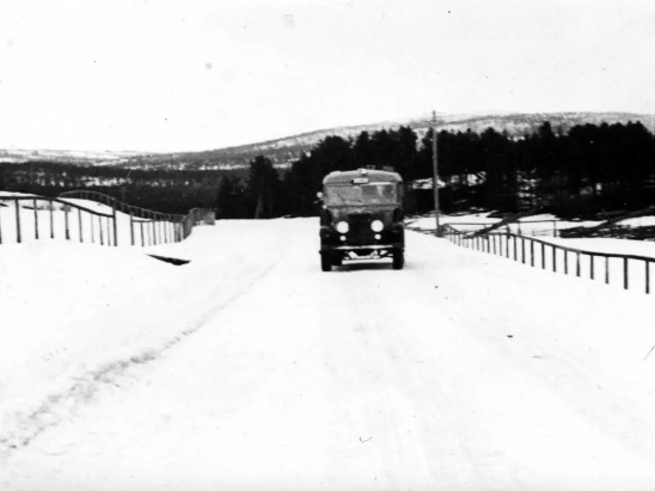 The image shows an old truck driving down a snowy road, surrounded by a fence on either side. In...