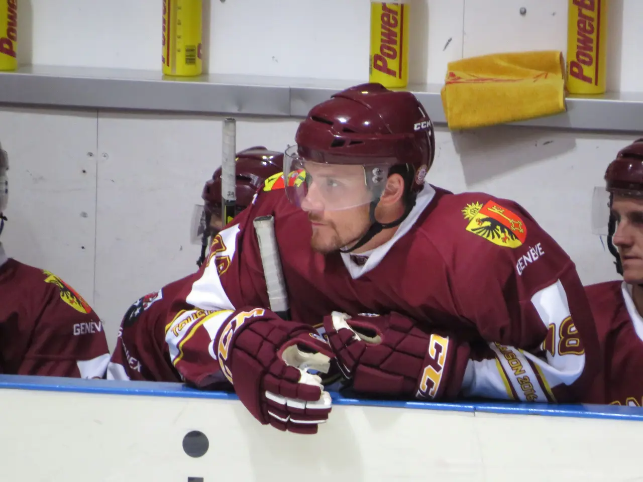 The image shows a group of hockey players sitting on top of a bench, wearing helmets and gloves. In...
