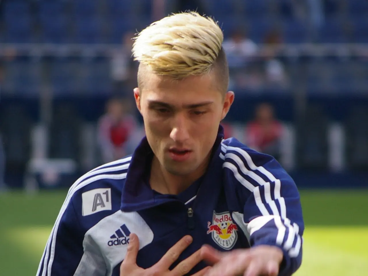 The image shows a young man in a blue t-shirt standing on a soccer field, with a blurred...