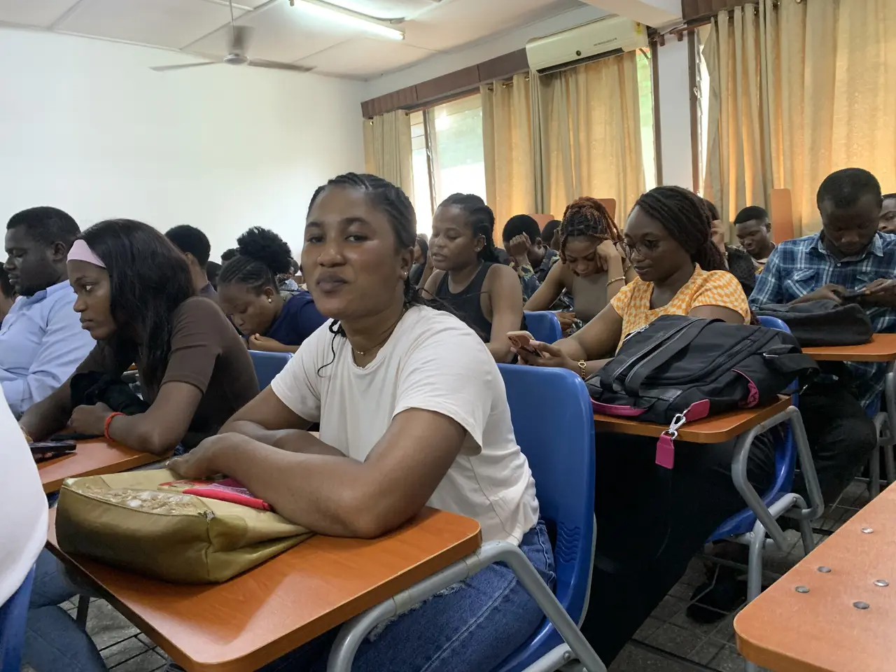 The image shows a group of people sitting at desks in a classroom, with bags and other objects on...