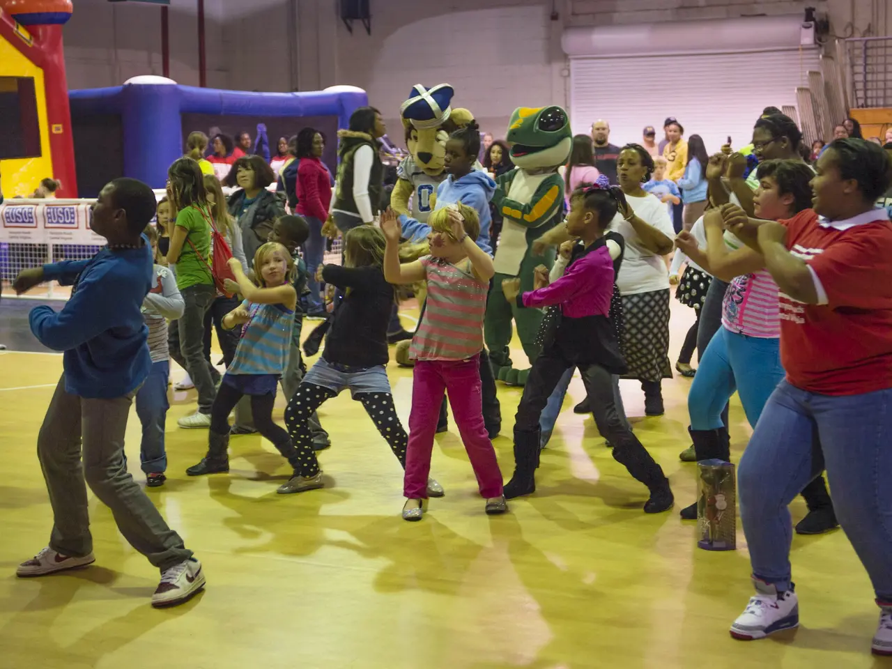 The image shows a group of children dancing in a gymnasium with a mascot in the center. There are...