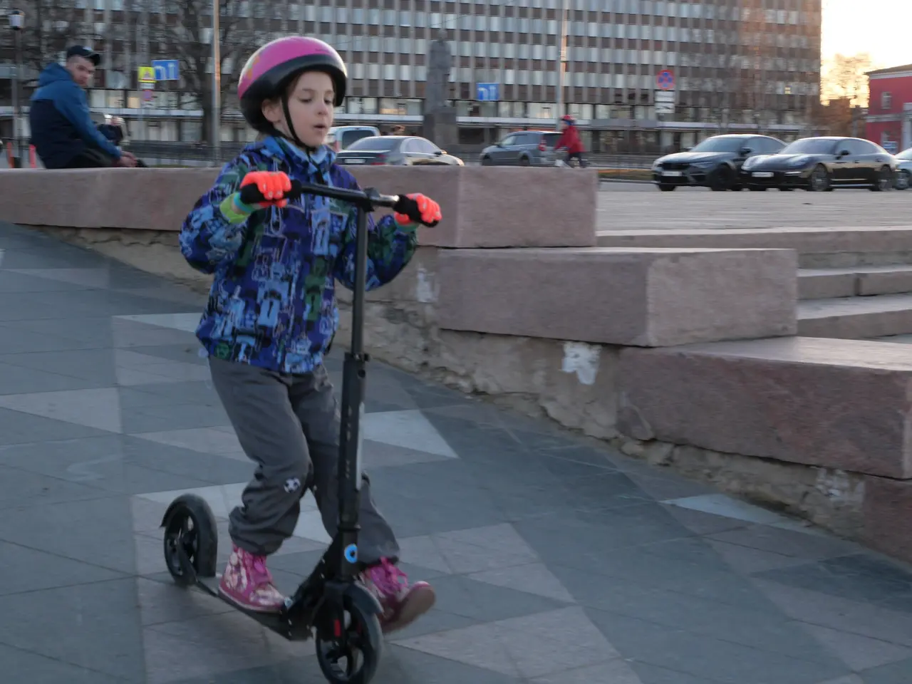 The image shows a young boy riding a scooter down a sidewalk, wearing a helmet and gloves. In the...