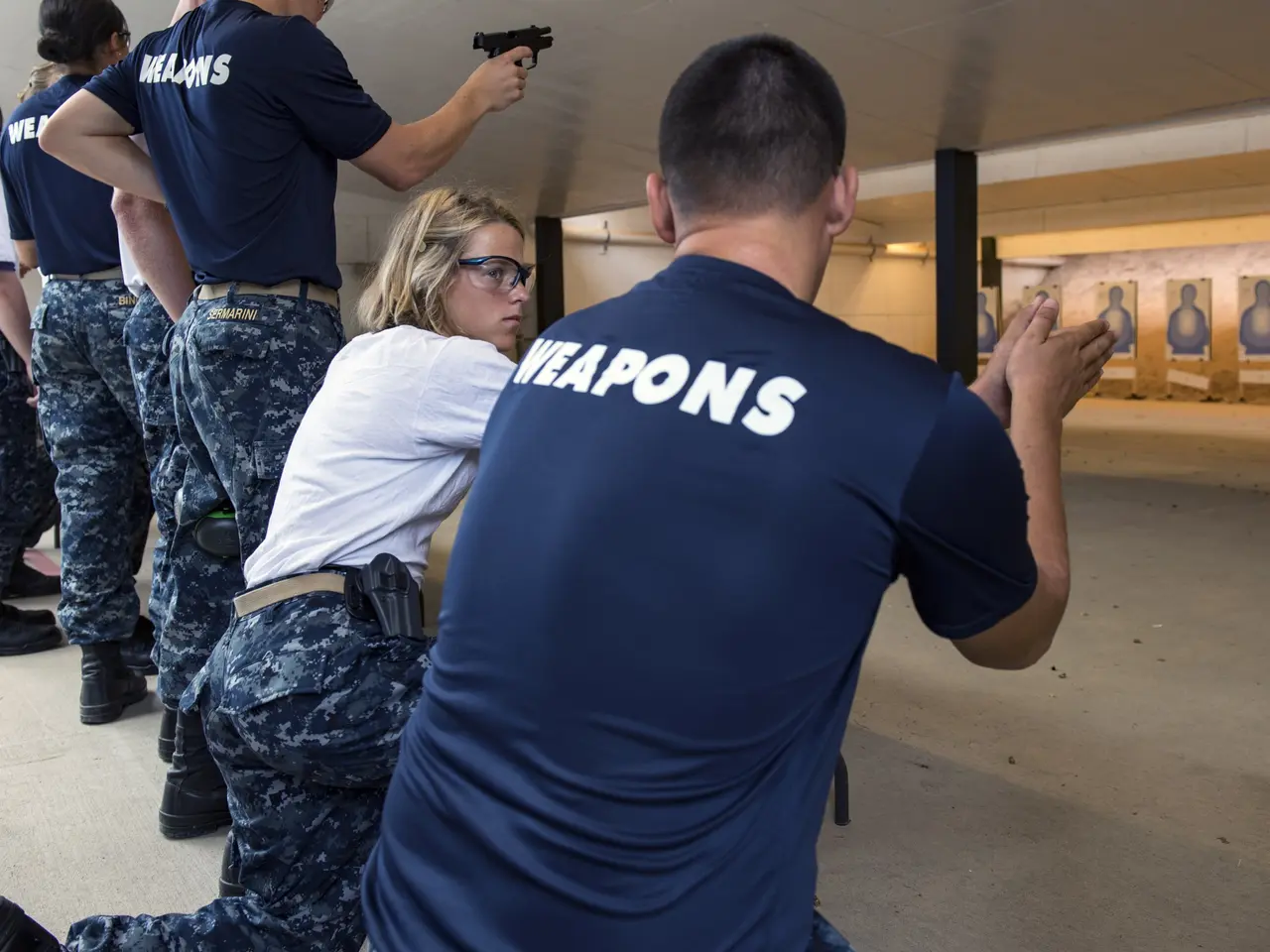 The image shows a group of people in blue shirts and black pants standing in a shooting range, with...