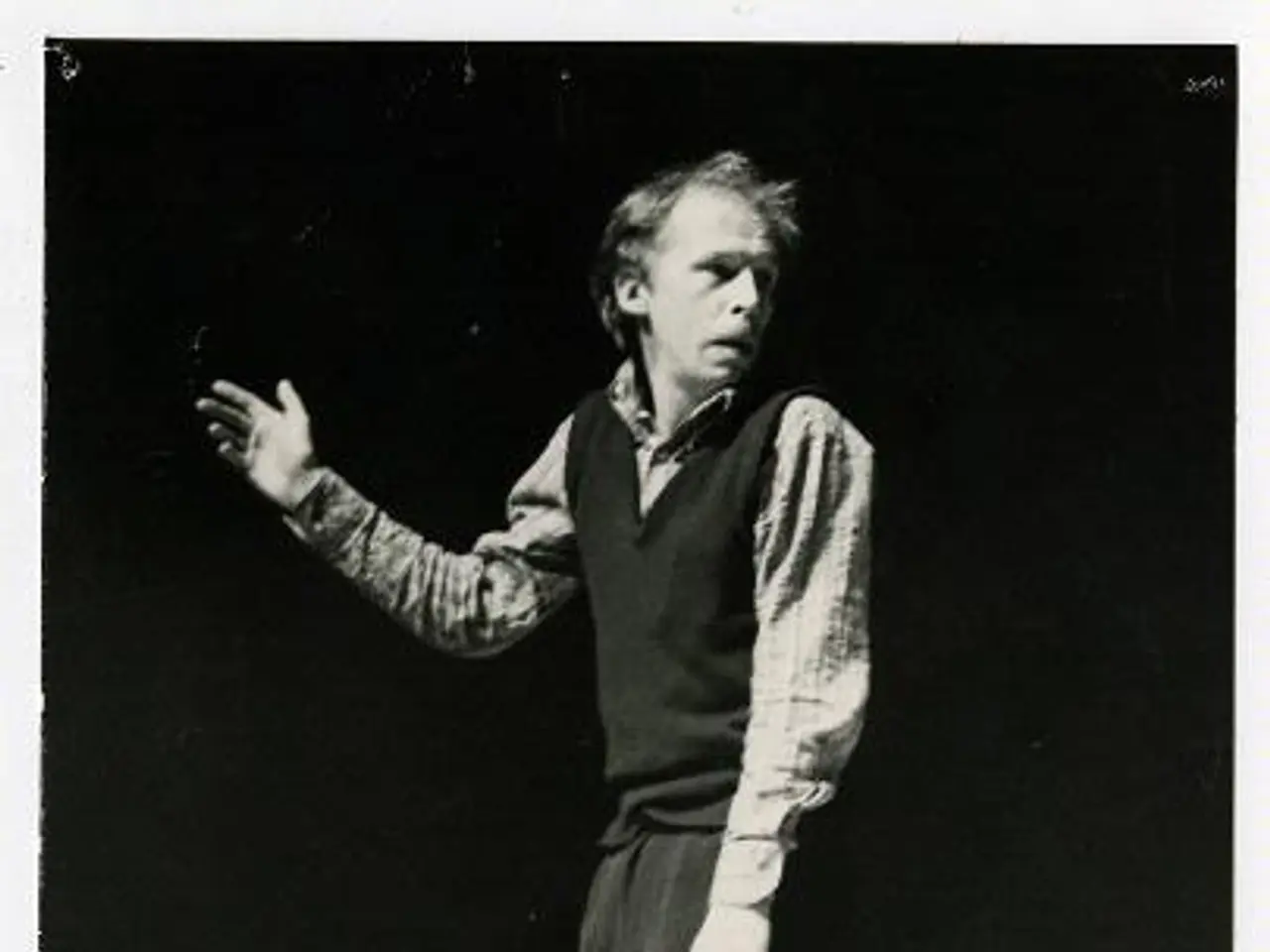 The image shows a man standing on a stage in a black and white photo. He is wearing a white shirt...