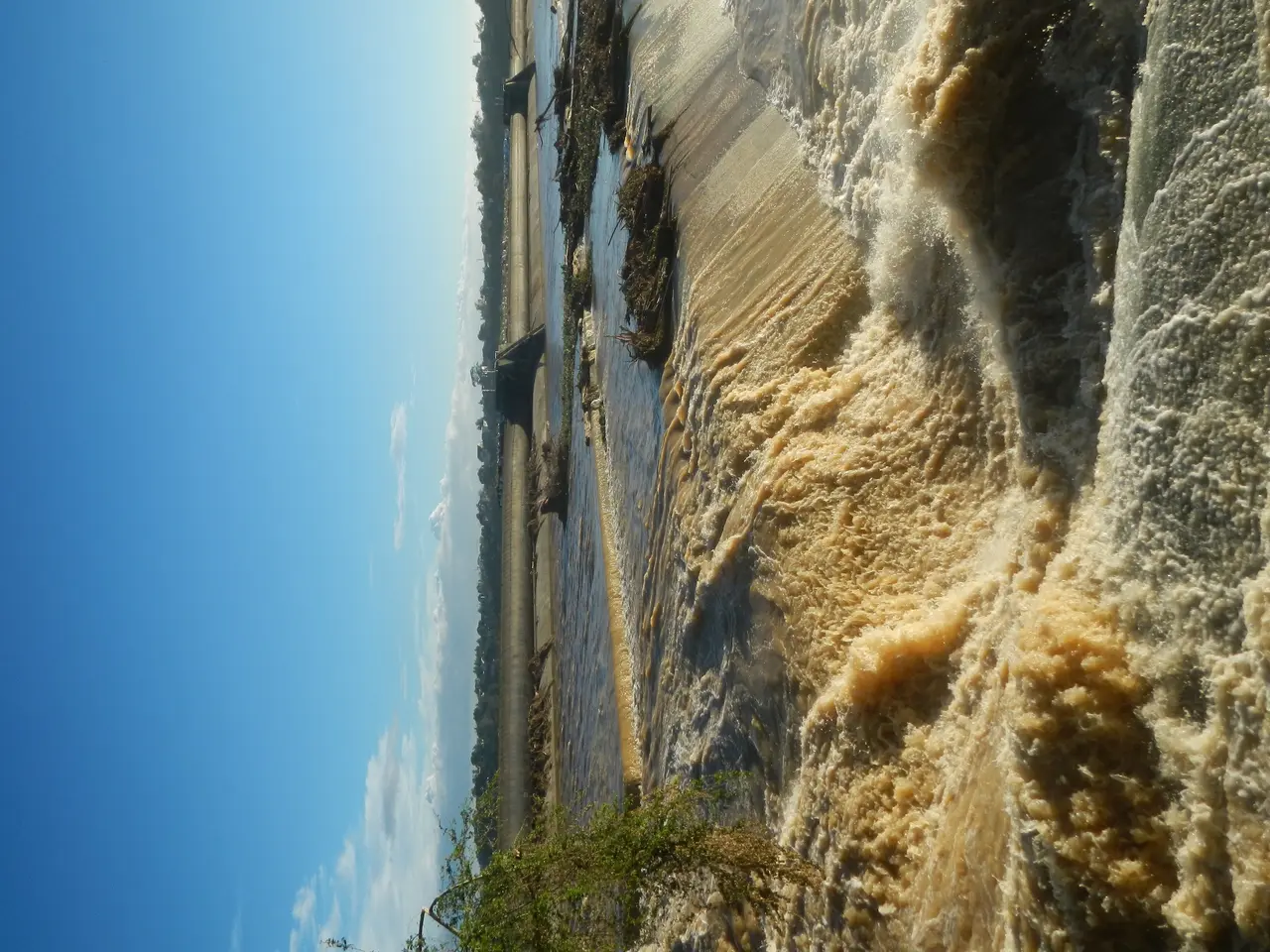 The image shows a dam with water flowing out of it, surrounded by lush green plants and a bright...