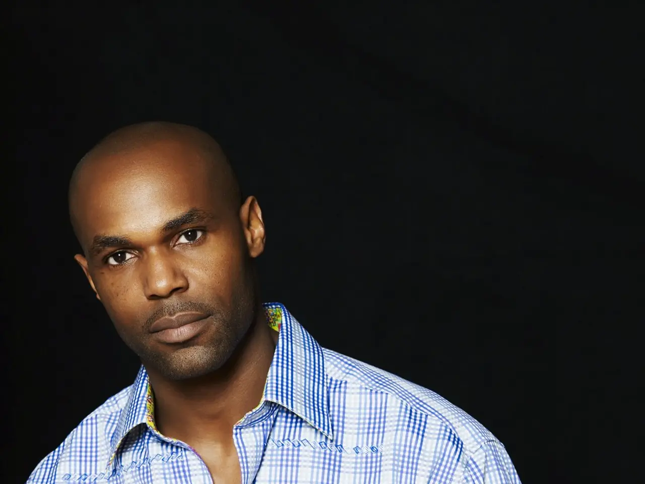 The image shows a man in a blue and white checkered shirt standing in front of a black background,...