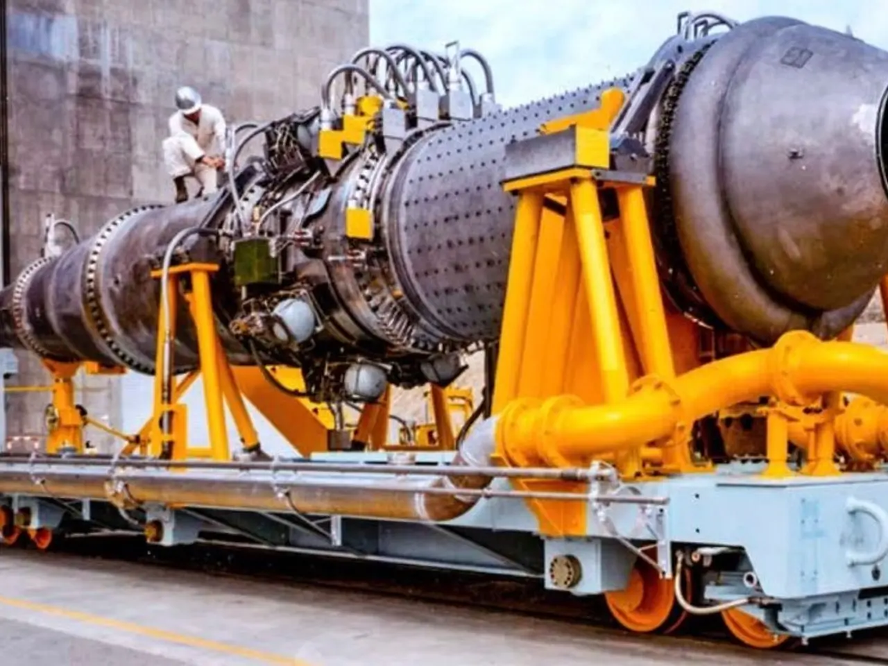 The image shows a large jet engine sitting atop a train on a track, with a person standing nearby....