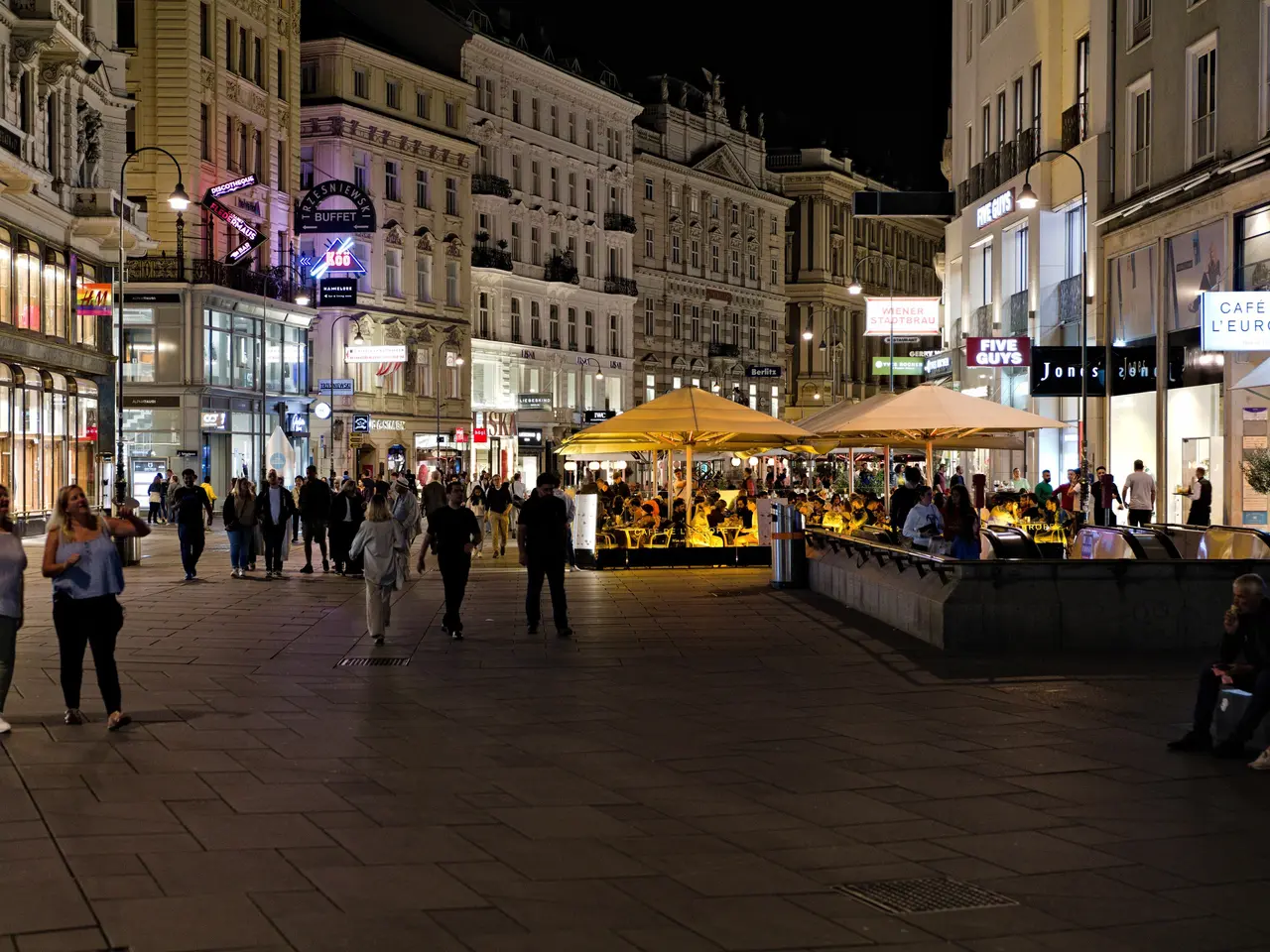 The image shows a bustling city street at night with people walking down it. There are buildings...