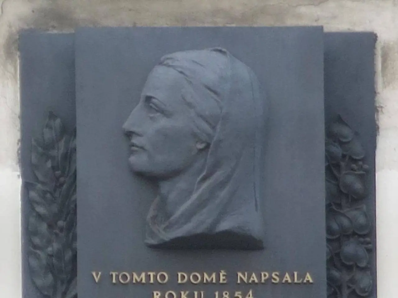 The image shows a plaque on the side of a building with a sculpture of a woman's face and text...
