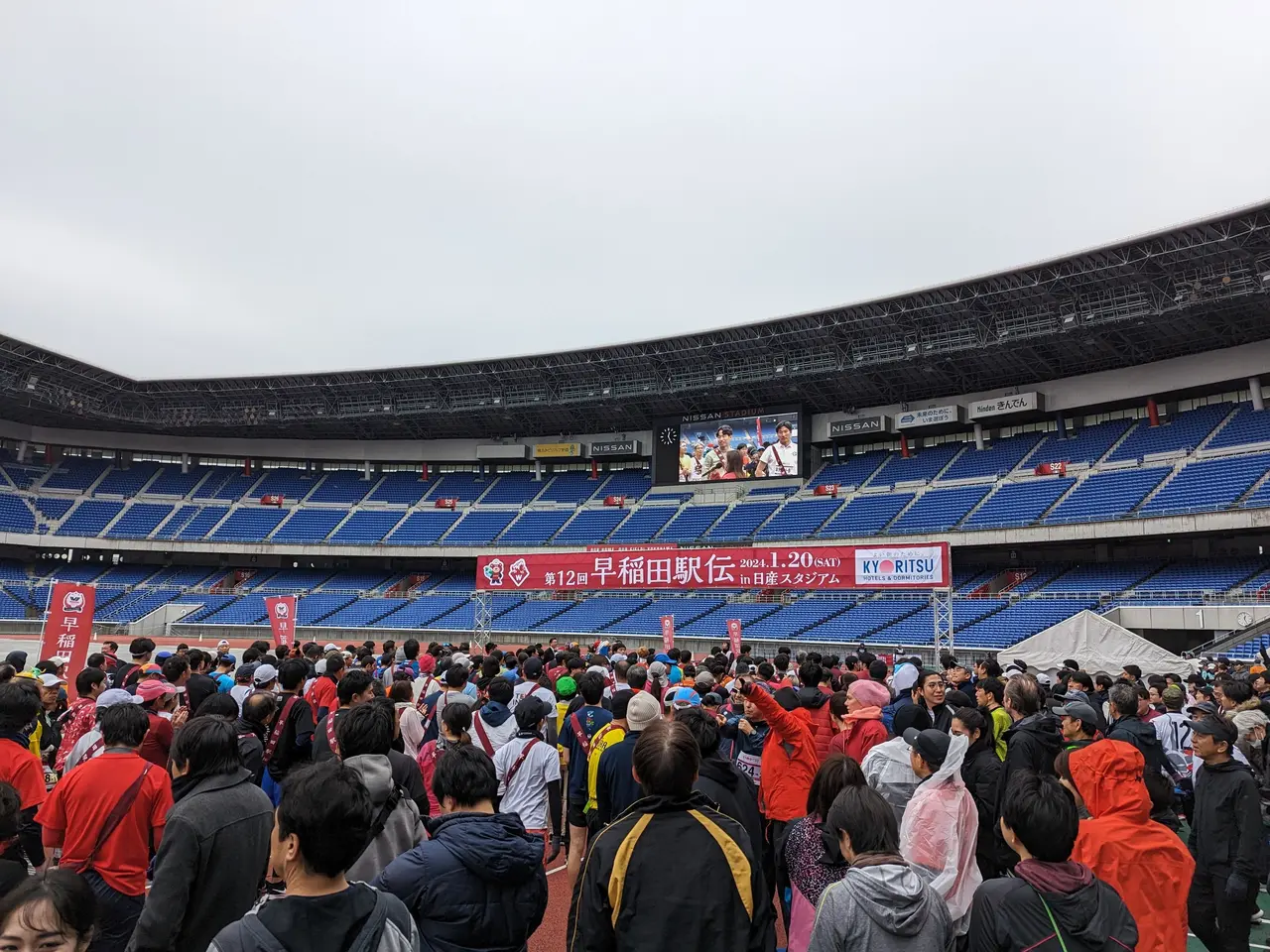 The image shows a large crowd of people standing in front of a stadium, with banners, chairs, and a...