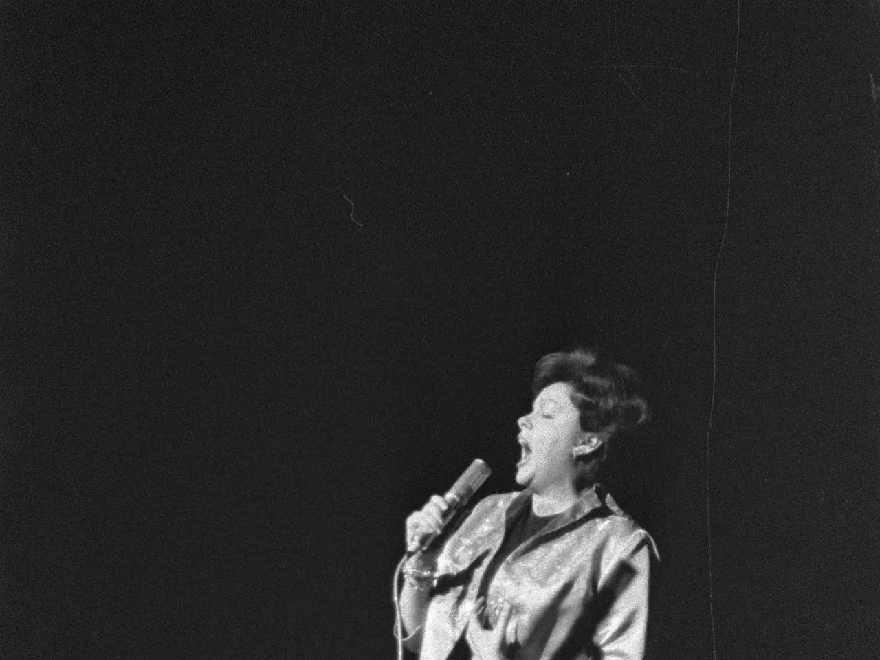 The image shows a woman standing on a stage, illuminated by a spotlight, singing into a microphone....