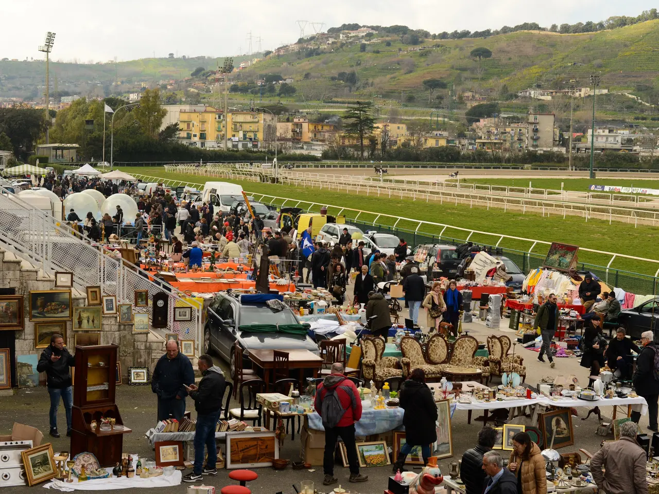 The image shows a large group of people standing around a flea market, with tables filled with...