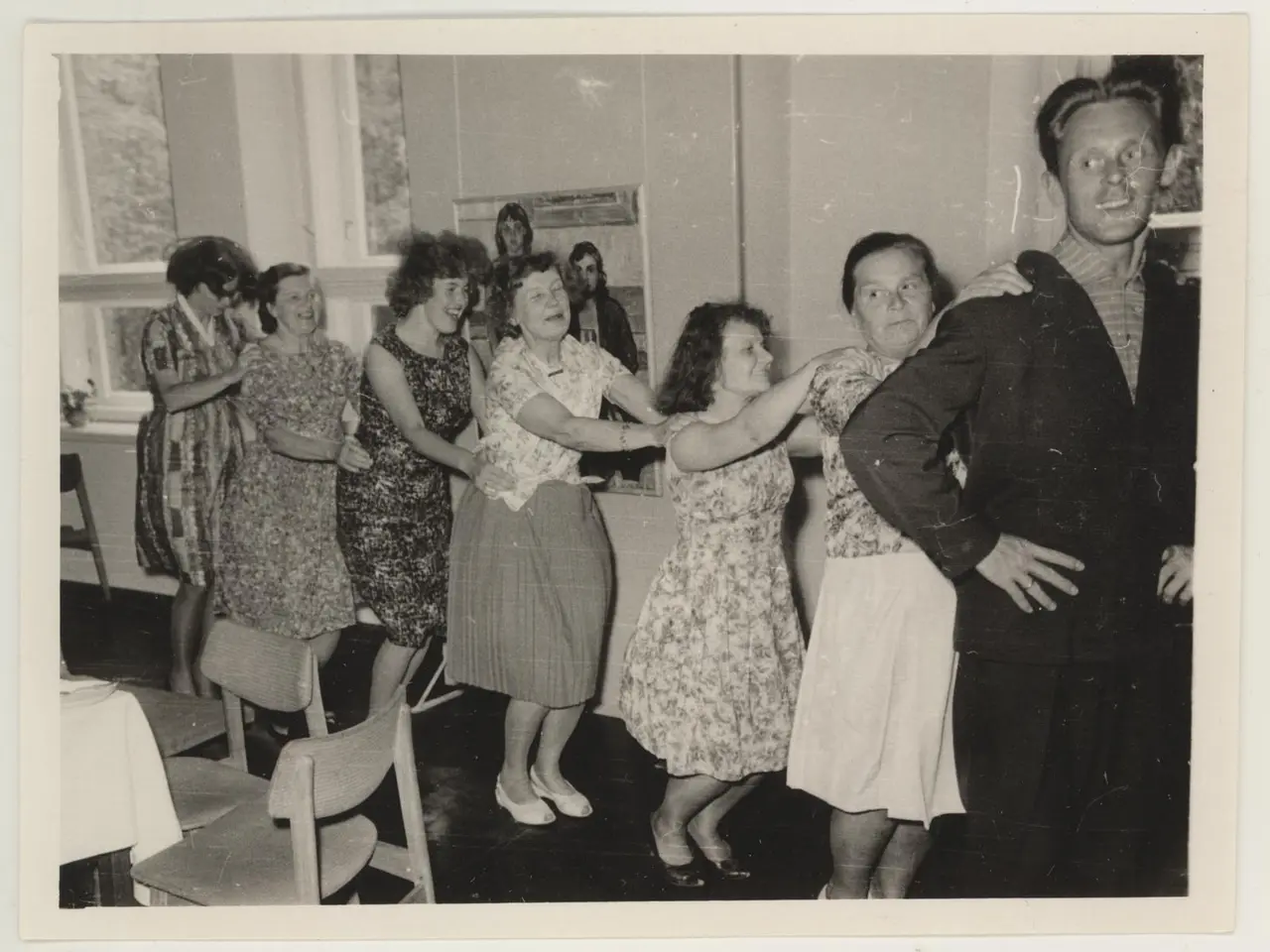 The image shows a group of people dancing in a room with chairs and a table covered with a white...