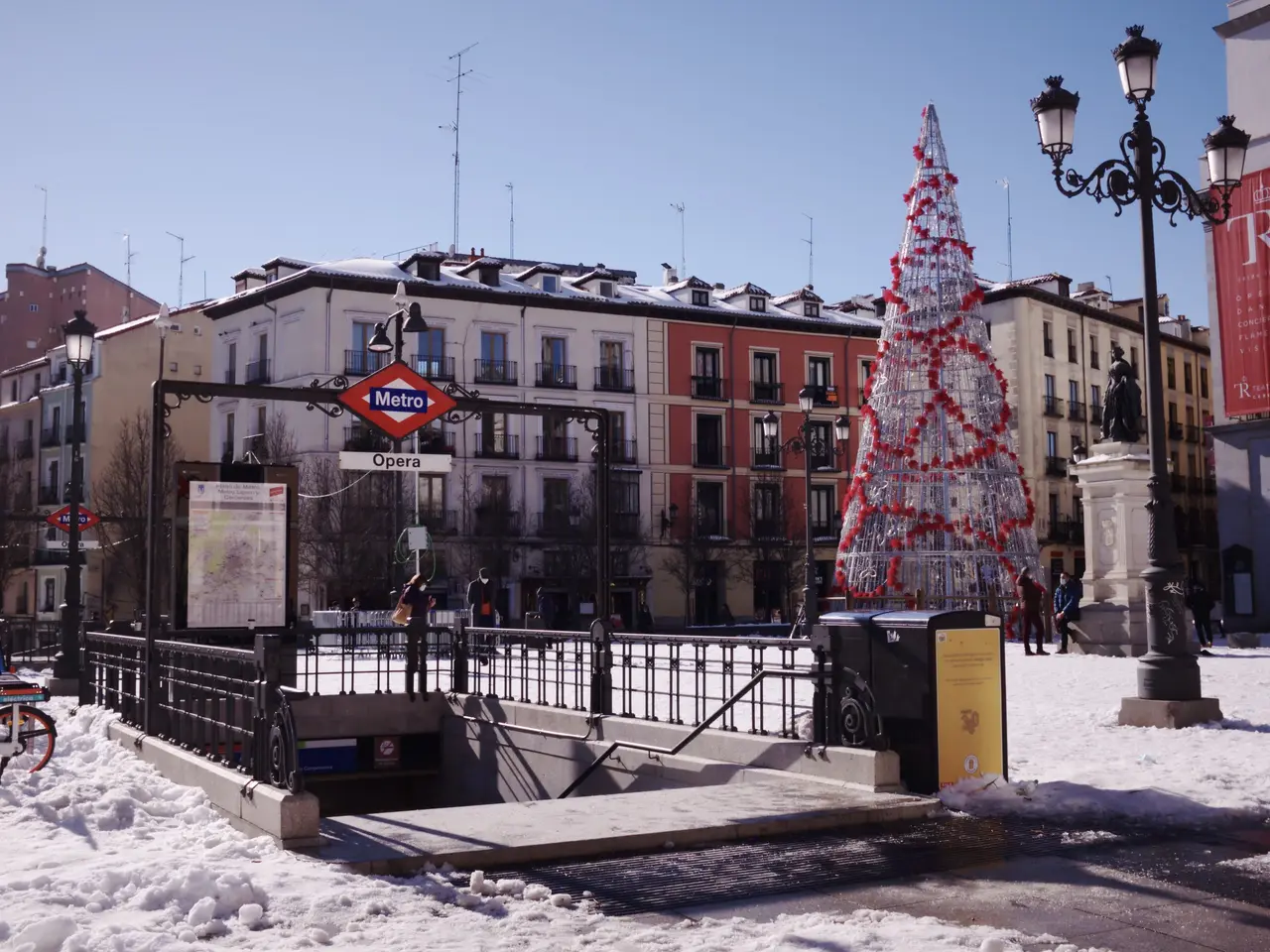 The image shows a Christmas tree in the middle of a snowy street, surrounded by buildings, street...