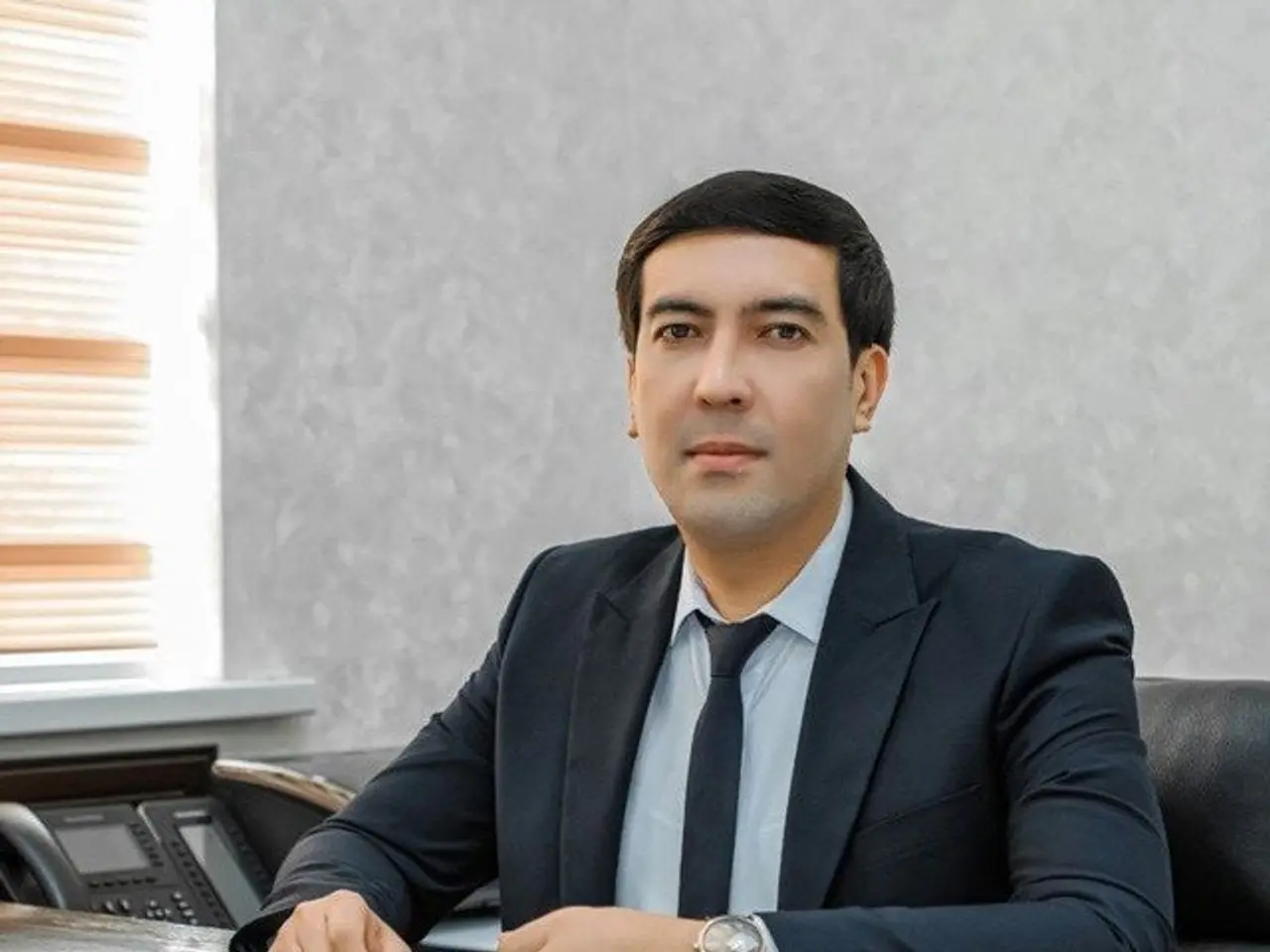 The image shows a man in a suit sitting at a desk with papers, a telephone, and other objects...