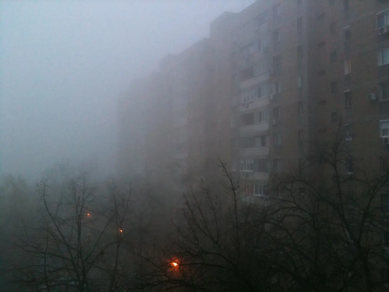 The image shows a foggy city street with tall buildings in the background, illuminated by street...