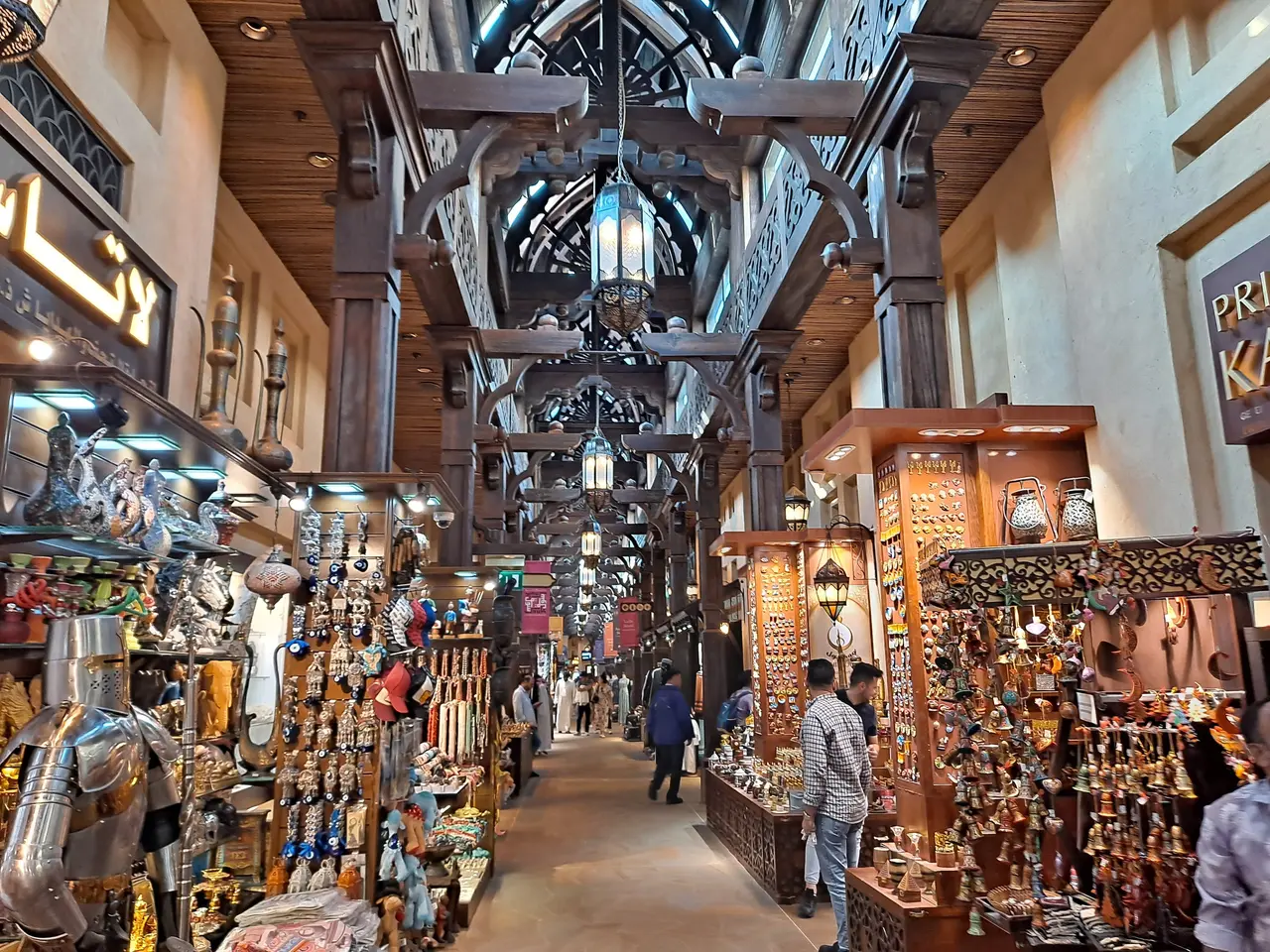 The image shows a bustling bazaar in Dubai, with people walking through it. On either side of the...