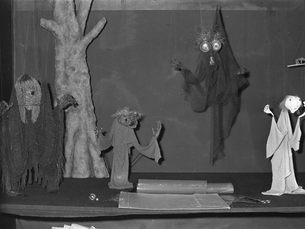 The image shows a black and white photo of a group of puppets on a table in front of a wall. On the...