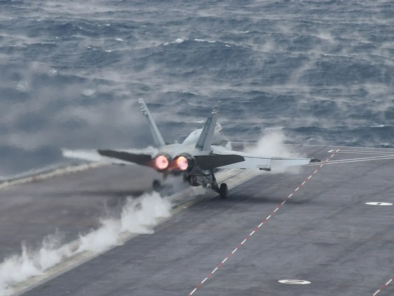The image shows a fighter jet taking off from an aircraft carrier in the middle of the ocean, with...