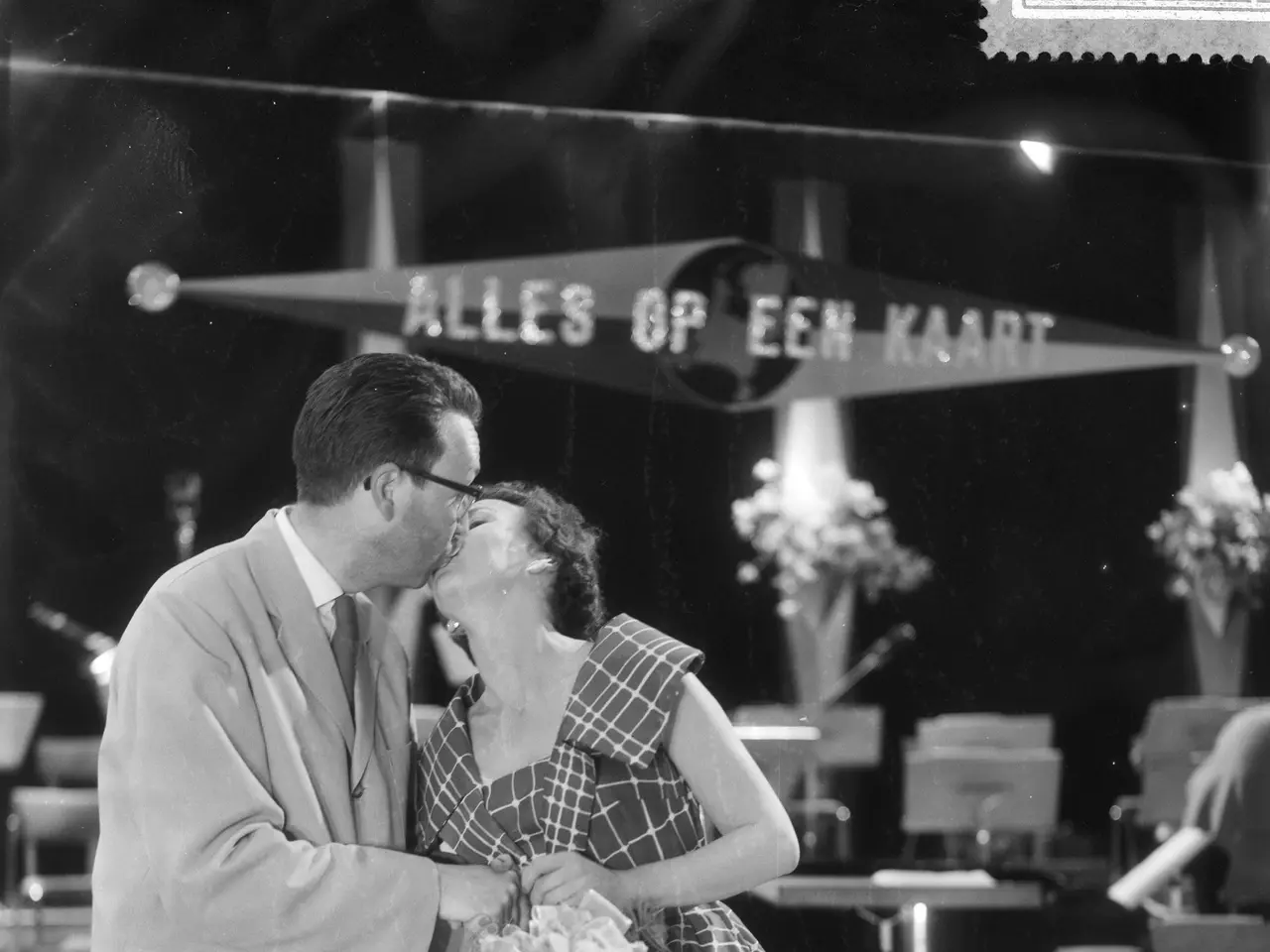 The image shows a man and woman embracing and kissing on a stage, with the woman holding a bouquet...