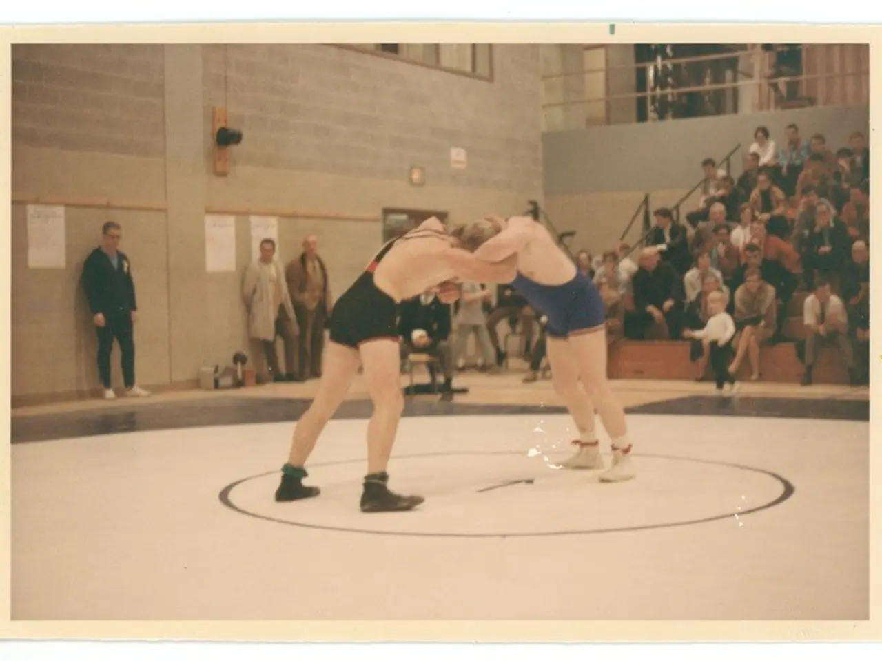 The image shows two wrestlers in the center of the frame, engaged in a wrestling match. Surrounding...