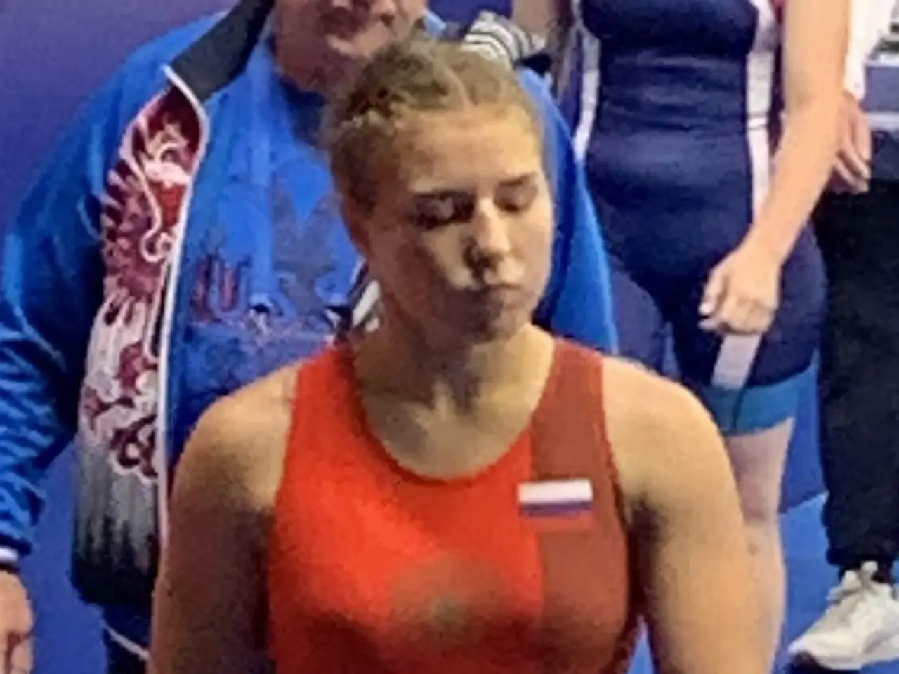 The image shows a woman in a red wrestling suit walking on a blue mat, surrounded by a group of...