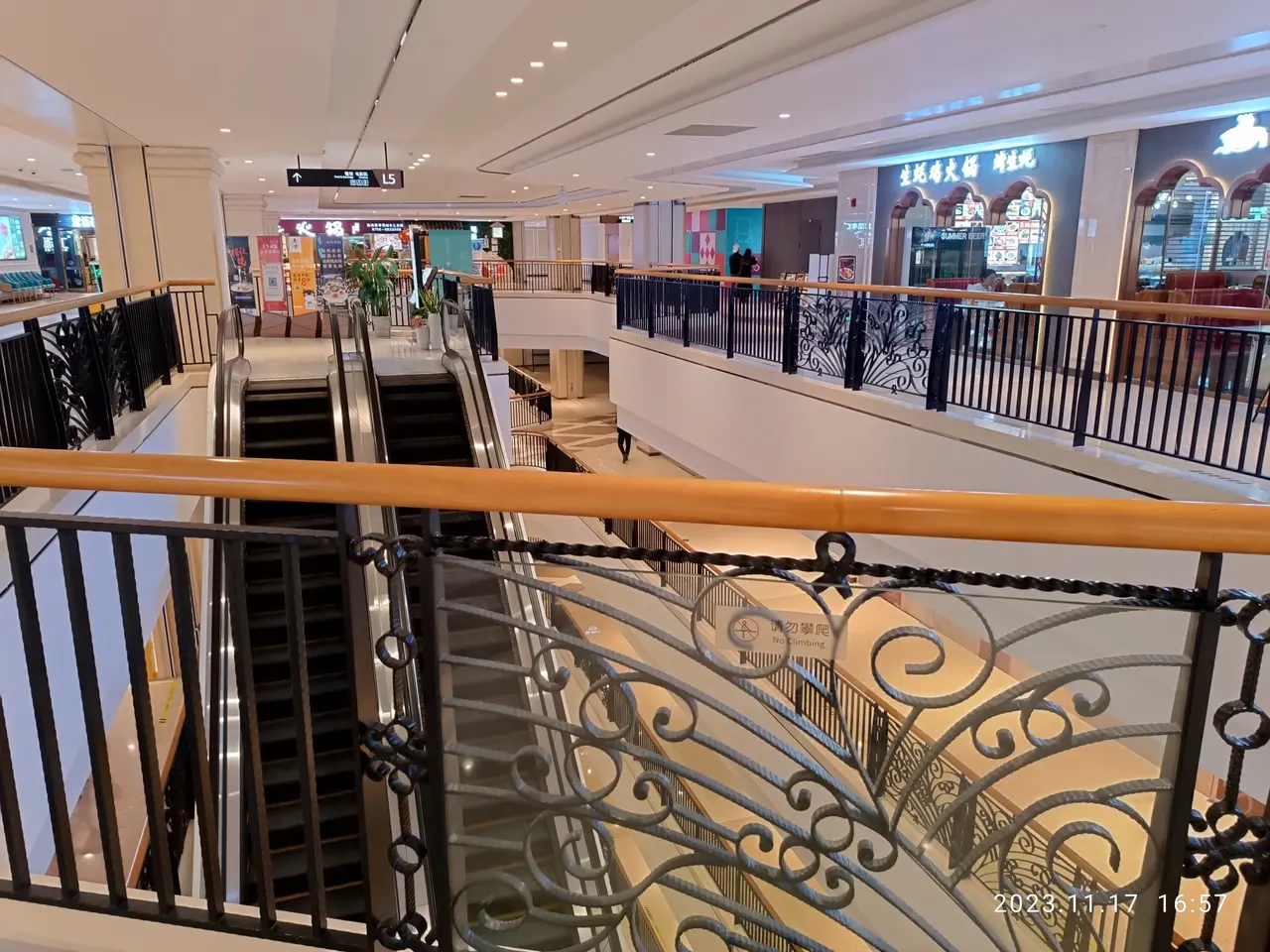 The image shows the interior of a shopping mall with escalators, railings, houseplants, stores,...