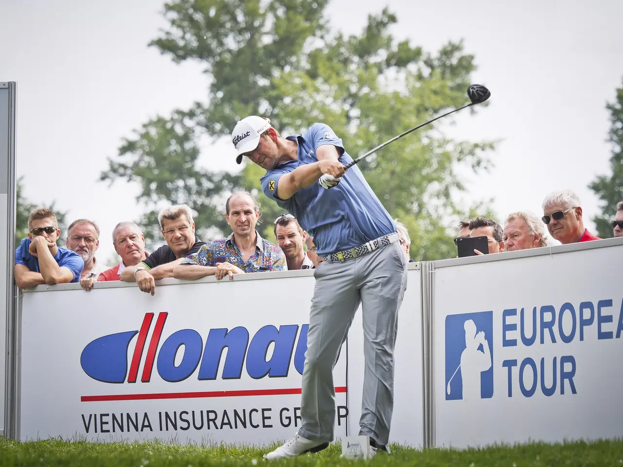 The image shows a man wearing a cap and swinging a golf club on top of a lush green field. In the...