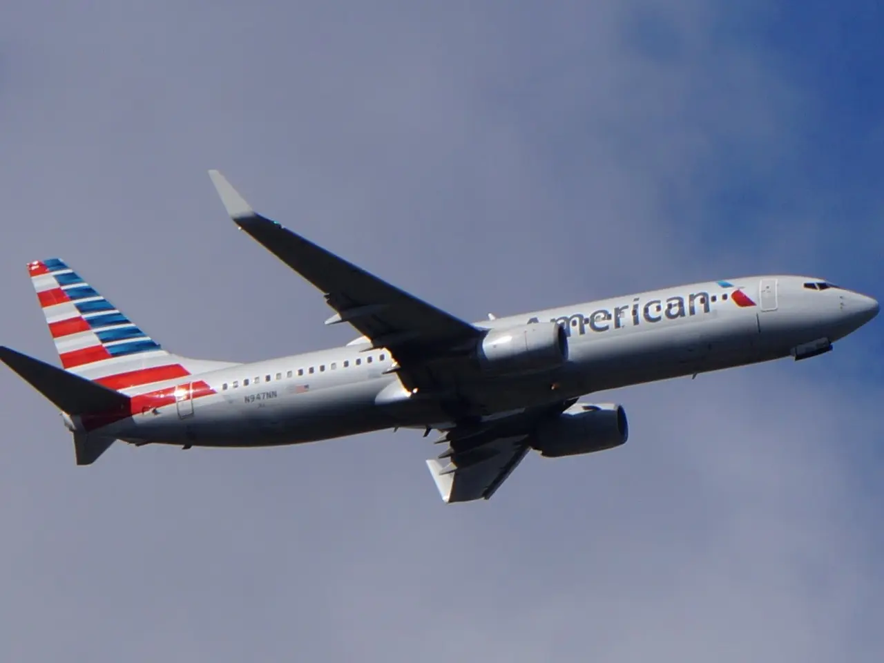 The image shows an American Airlines plane flying through a blue sky with white, fluffy clouds in...