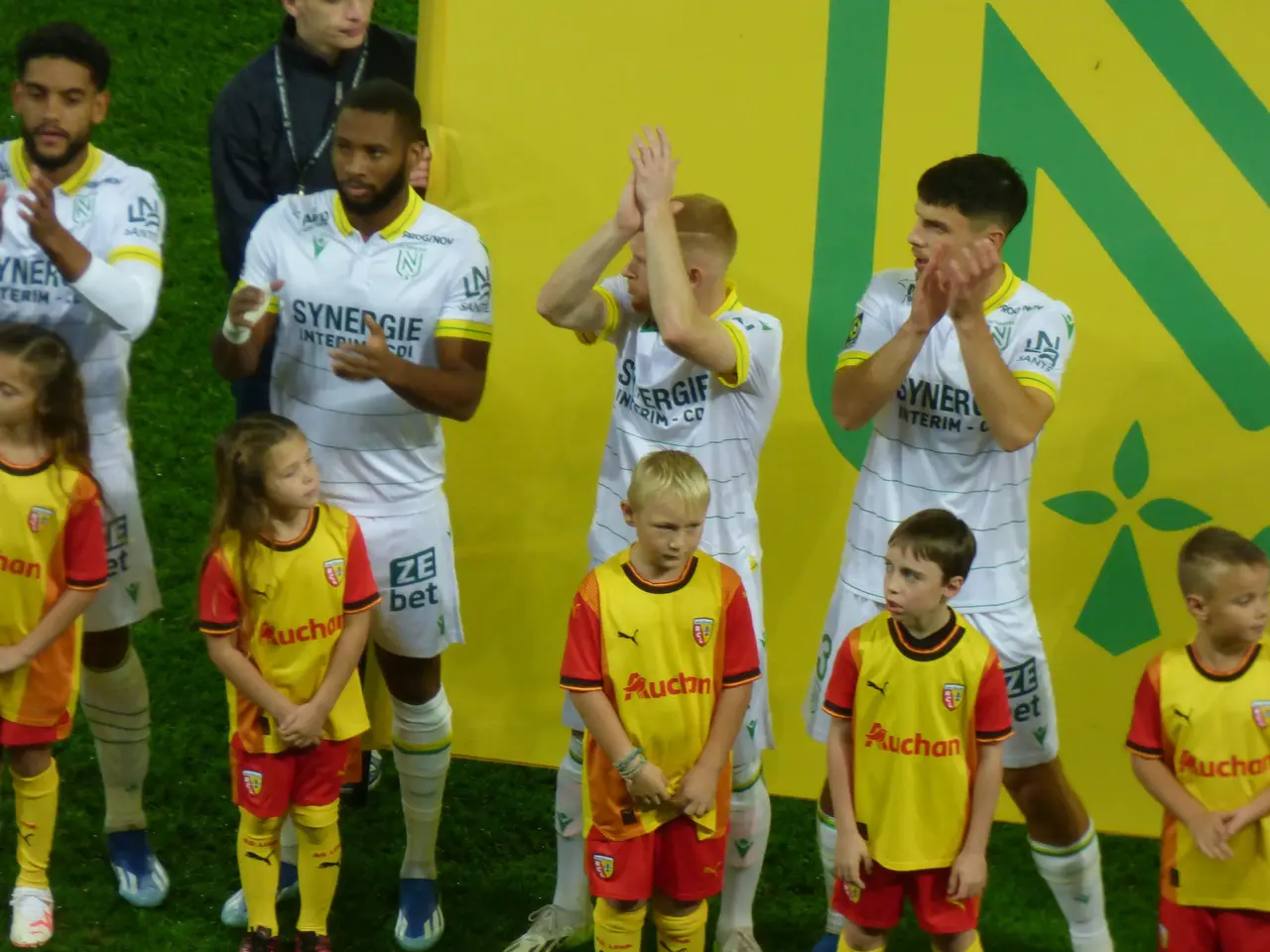The image shows a group of young people standing on top of a soccer field, clapping their hands in...