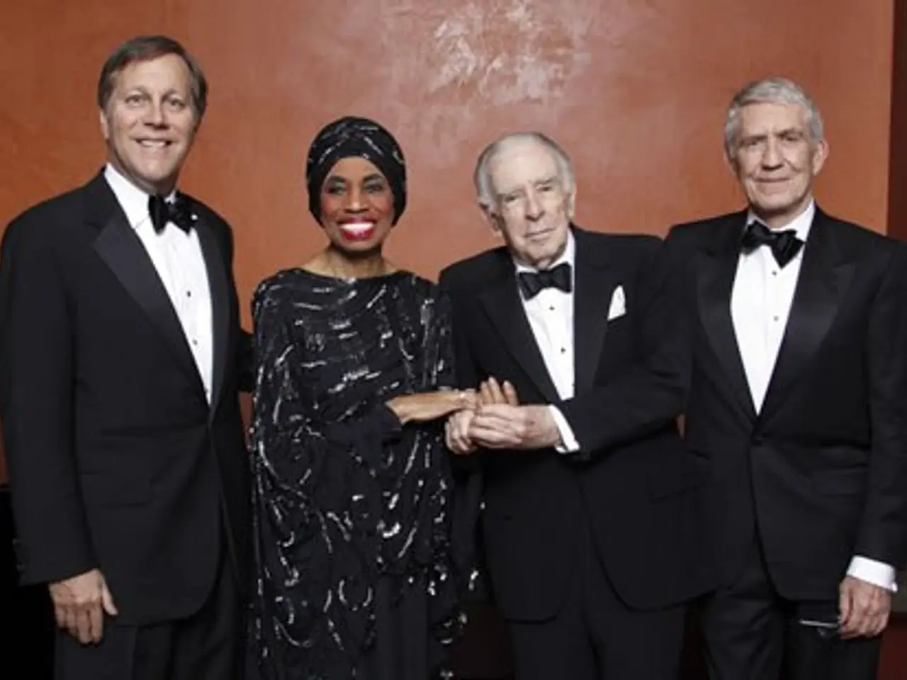 The image shows a group of four people in tuxedos standing next to each other, smiling and posing...