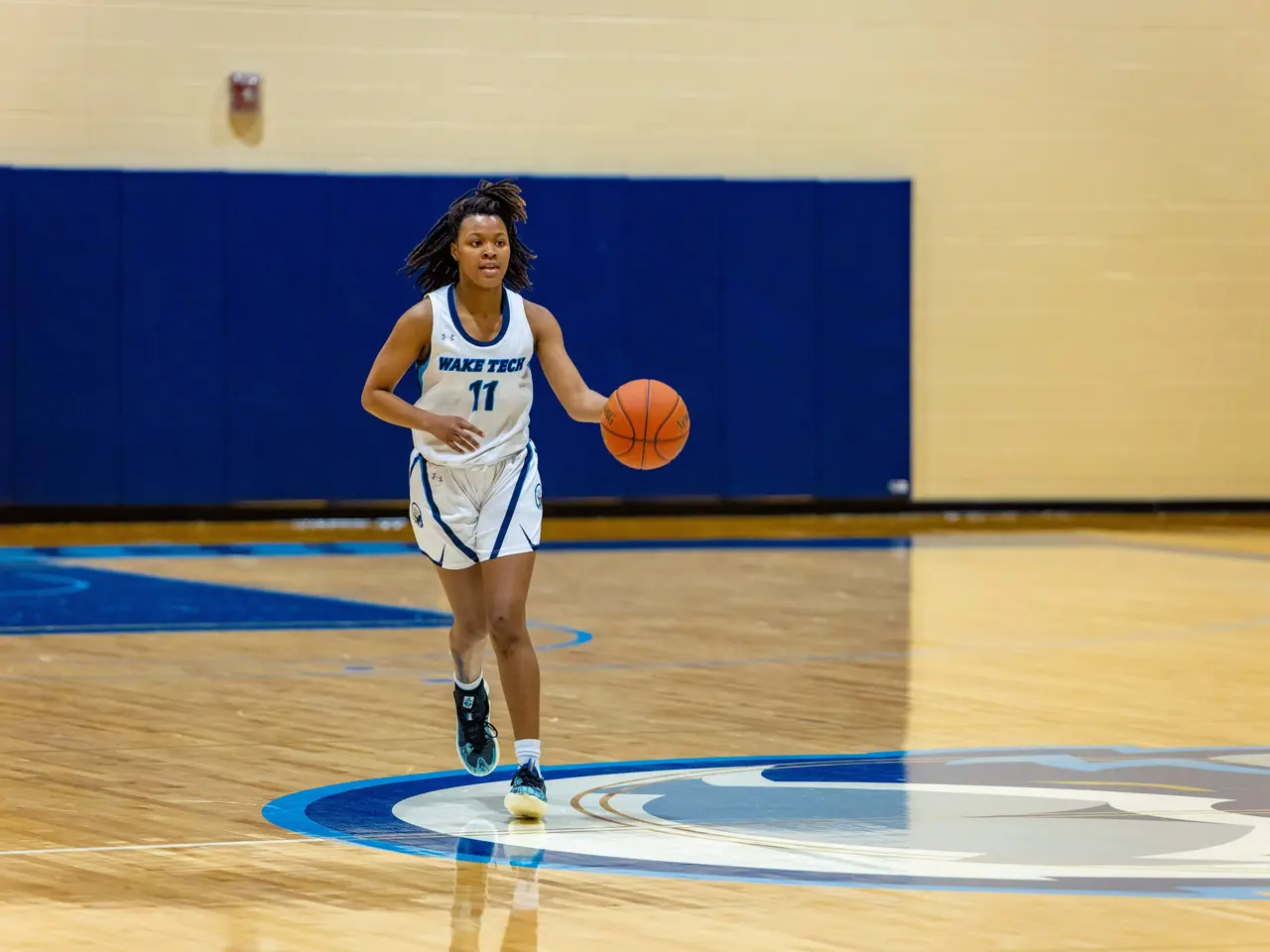 The image shows a woman in a blue and white uniform dribbling a basketball on a court, with a wall...