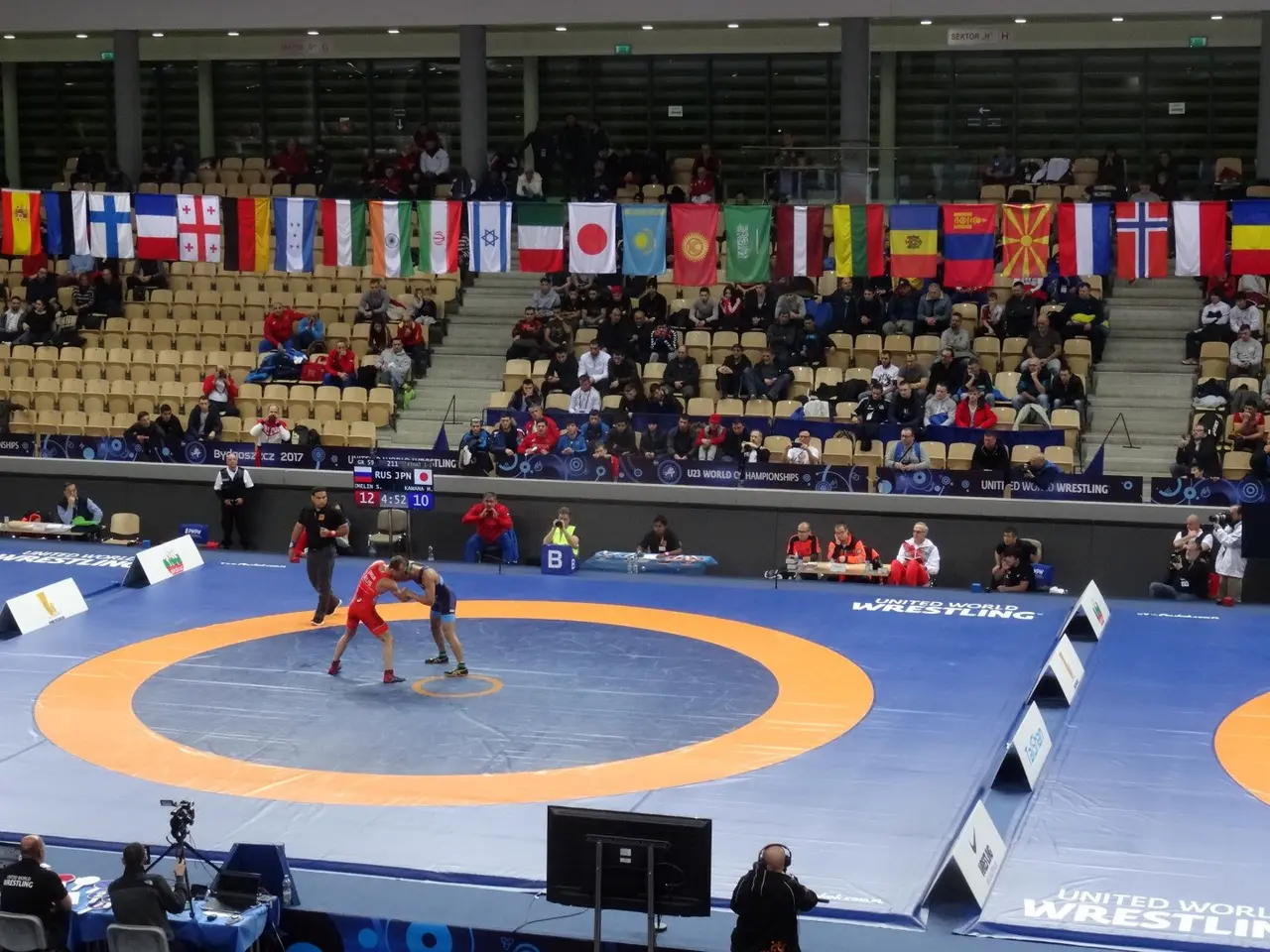 The image shows two wrestlers in the middle of a wrestling match in a large arena, surrounded by a...