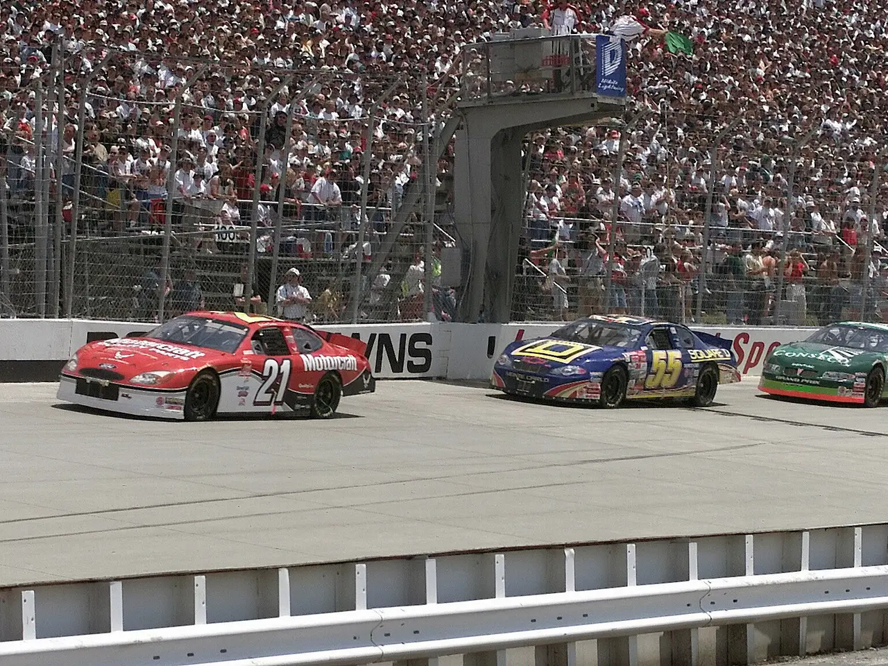 The image shows three NASCAR cars racing down a race track in front of a large crowd. The cars are...