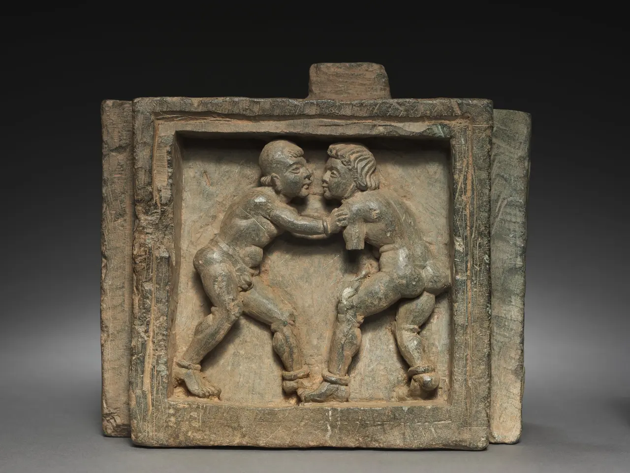 The image shows a terracotta plaque depicting two men engaged in a wrestling match. The plaque is...