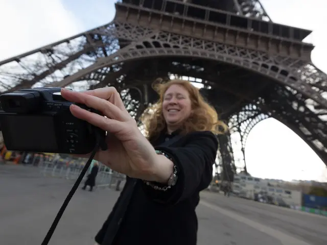 In this image I see a woman, who is smiling and holding a camera. In the background i see few...