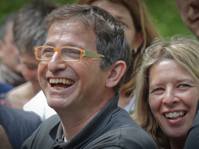 In the center of the image we can see a man wearing spectacles. We can also see a group of people...