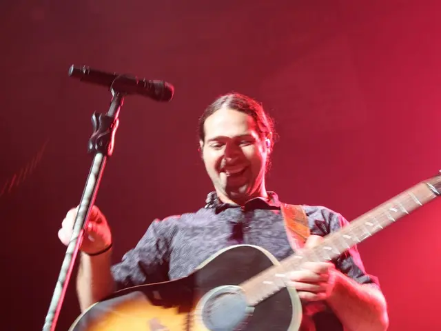 This man is holding a guitar and mic holder with mic. This man holds smile.