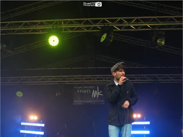 There are 2 UK flags hanging. A person is standing on the stage wearing a cap, black shirt and...