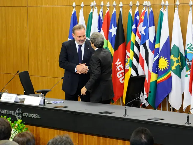 In this image we can see there are two people shaking hands and are standing behind a table, which...