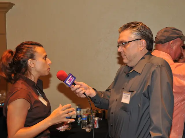 In this picture a guy is holding a mic in his hand and interviewing a lady. To the right side of...