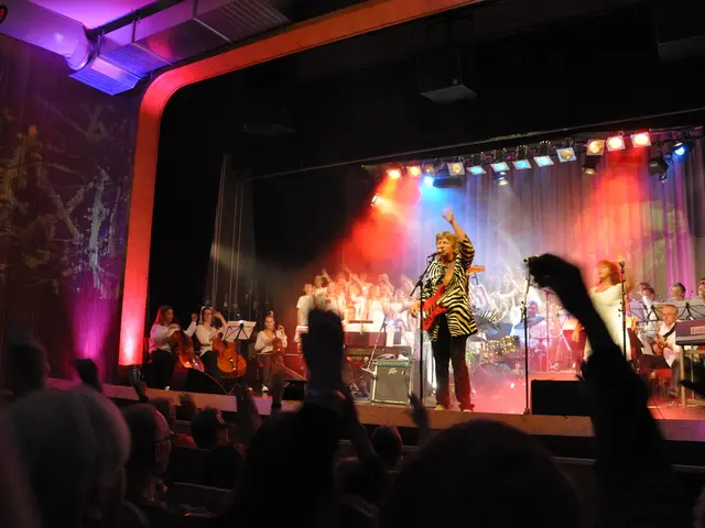 In this image at front people are sitting on the chairs. In front of them there is a stage. On a...